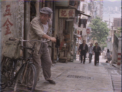 An elderly man holds his bicycle on a steep street while three men approach